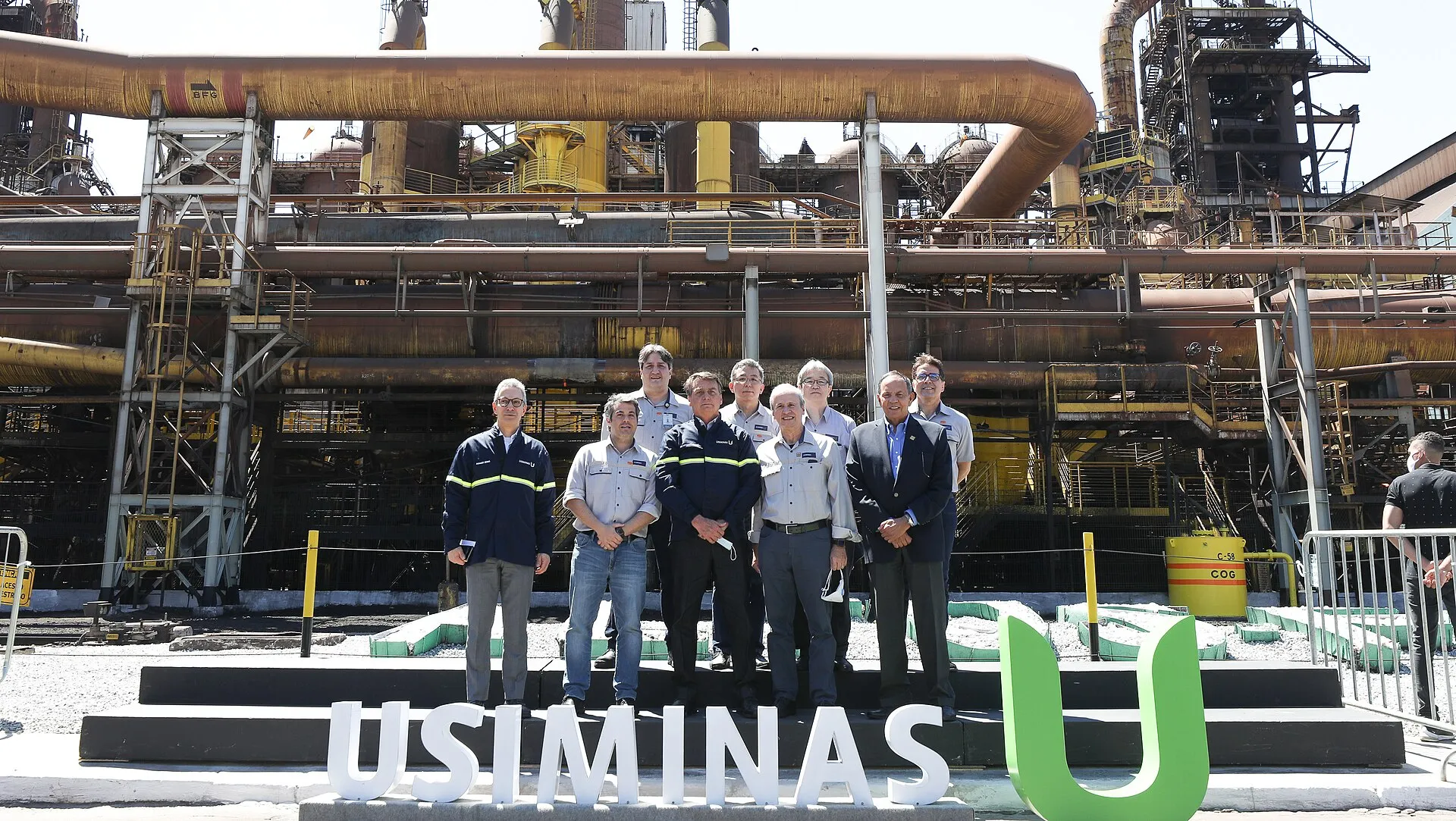 Grupo de homens posa em frente ao alto-forno 1 da usina da Usiminas, em Ipatinga, com tubulações industriais ao fundo e o logotipo da empresa em primeiro plano.