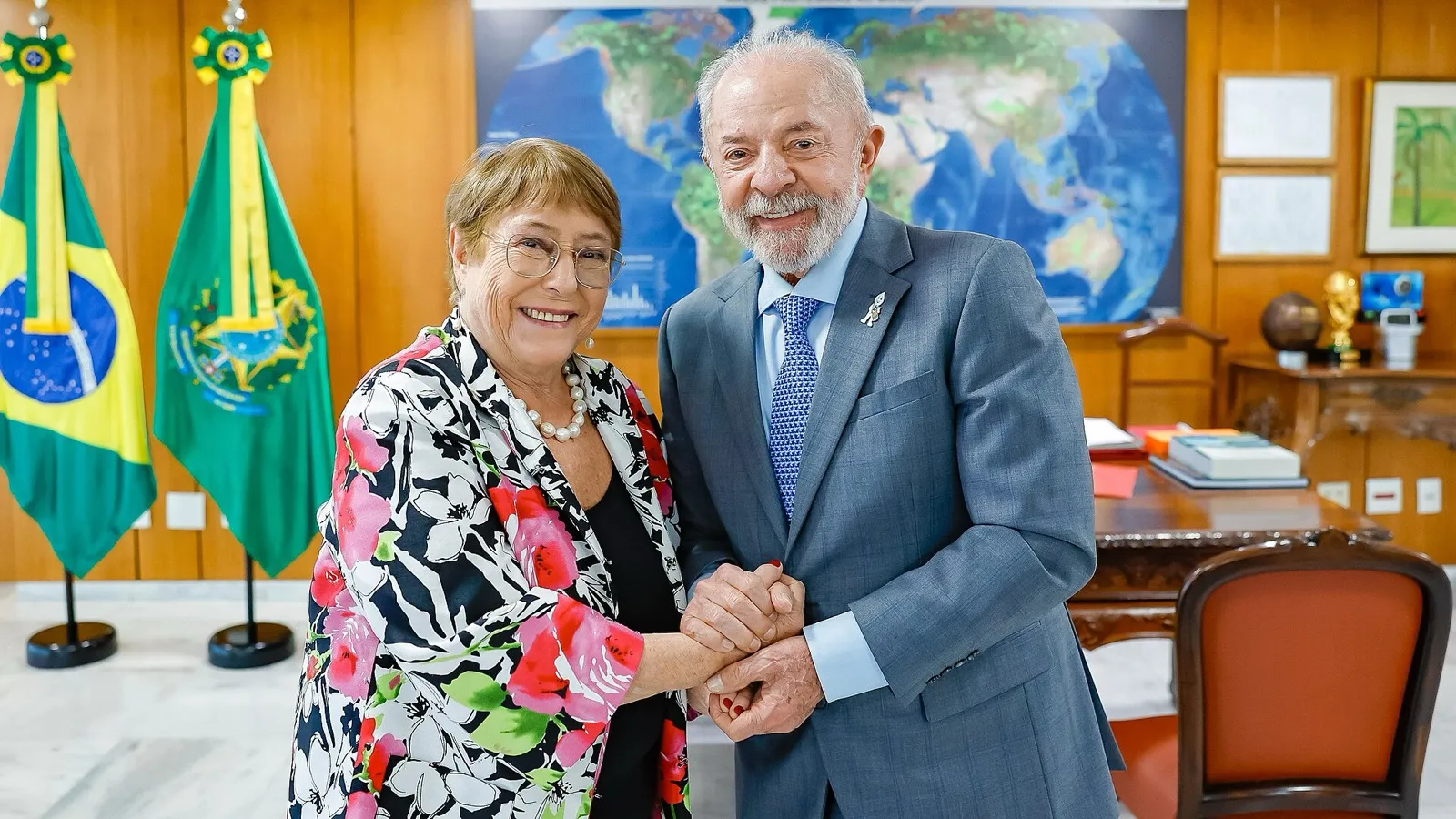 Lula e Michelle Bachelet, de mãos dadas, em uma sala do Palácio do Planalto, com as bandeiras do Brasil e da Presidência ao fundo.