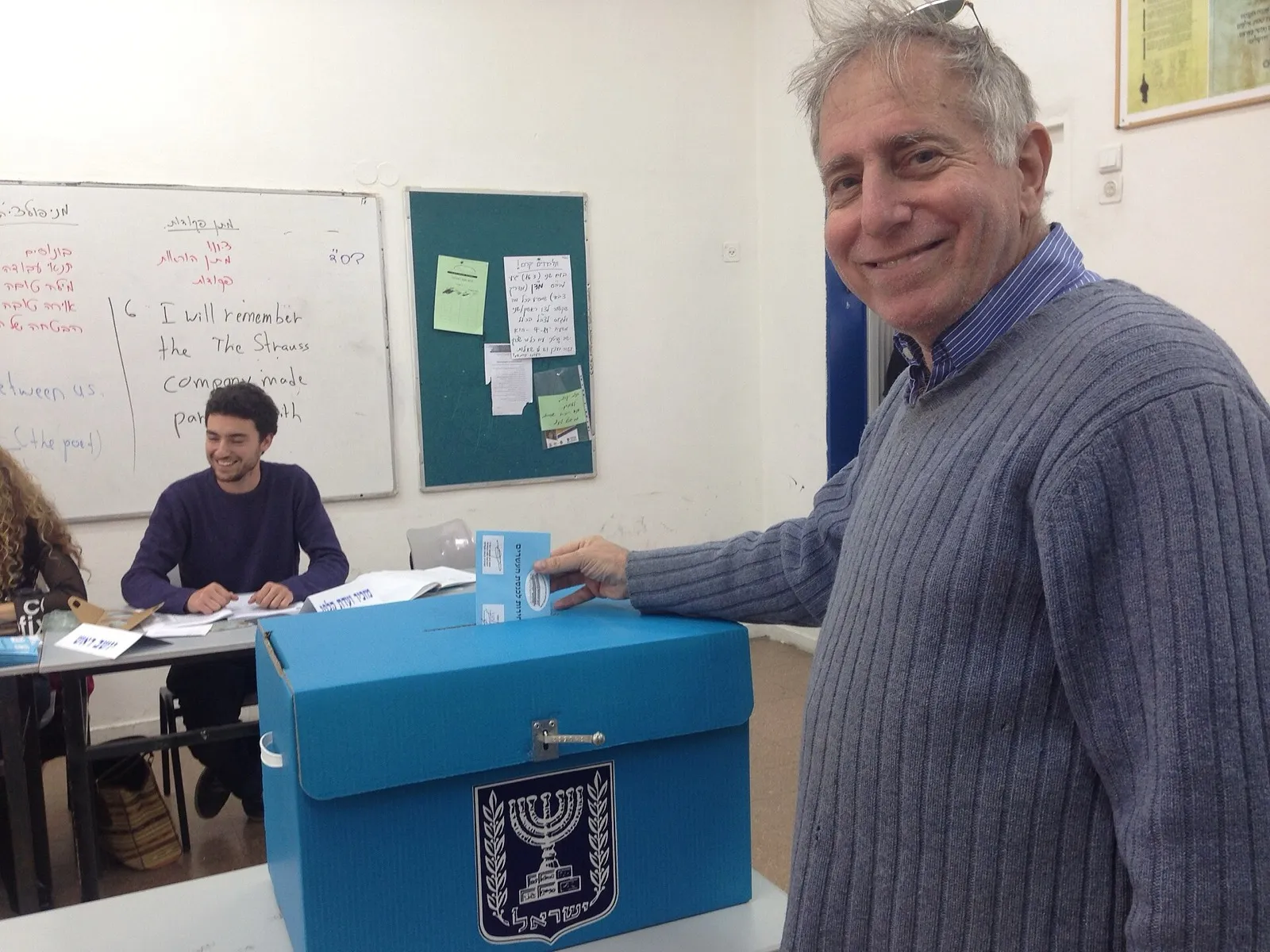 Eleitor deposita cédula azul em urna com o brasão de Israel durante votação em uma sala.