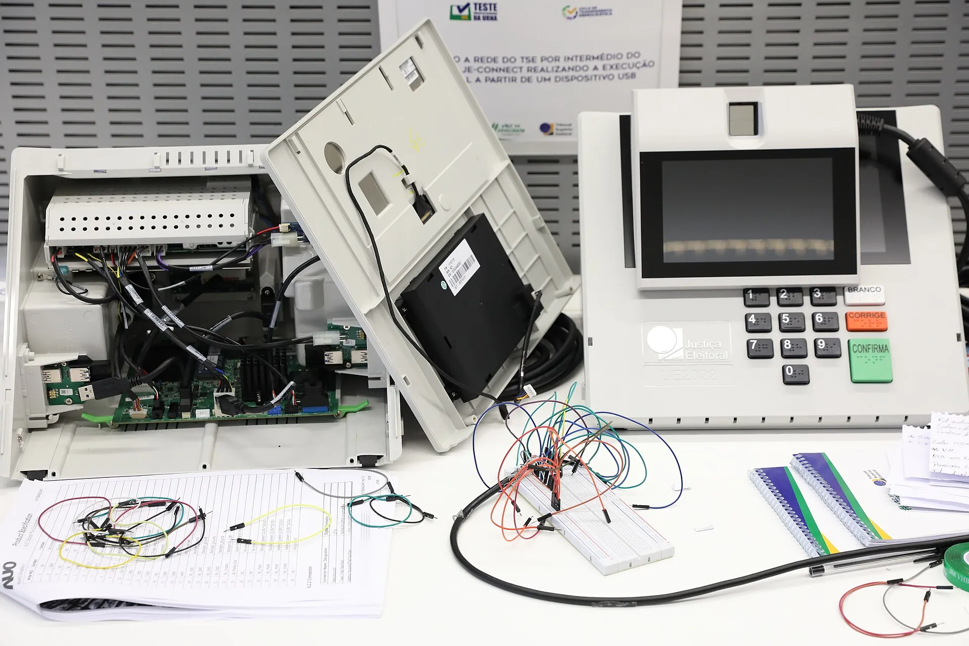 Urna eletrônica modelo UE2022 com tela e teclado intactos, ao lado de outra com a tampa aberta e placas expostas, sobre uma bancada com fios.