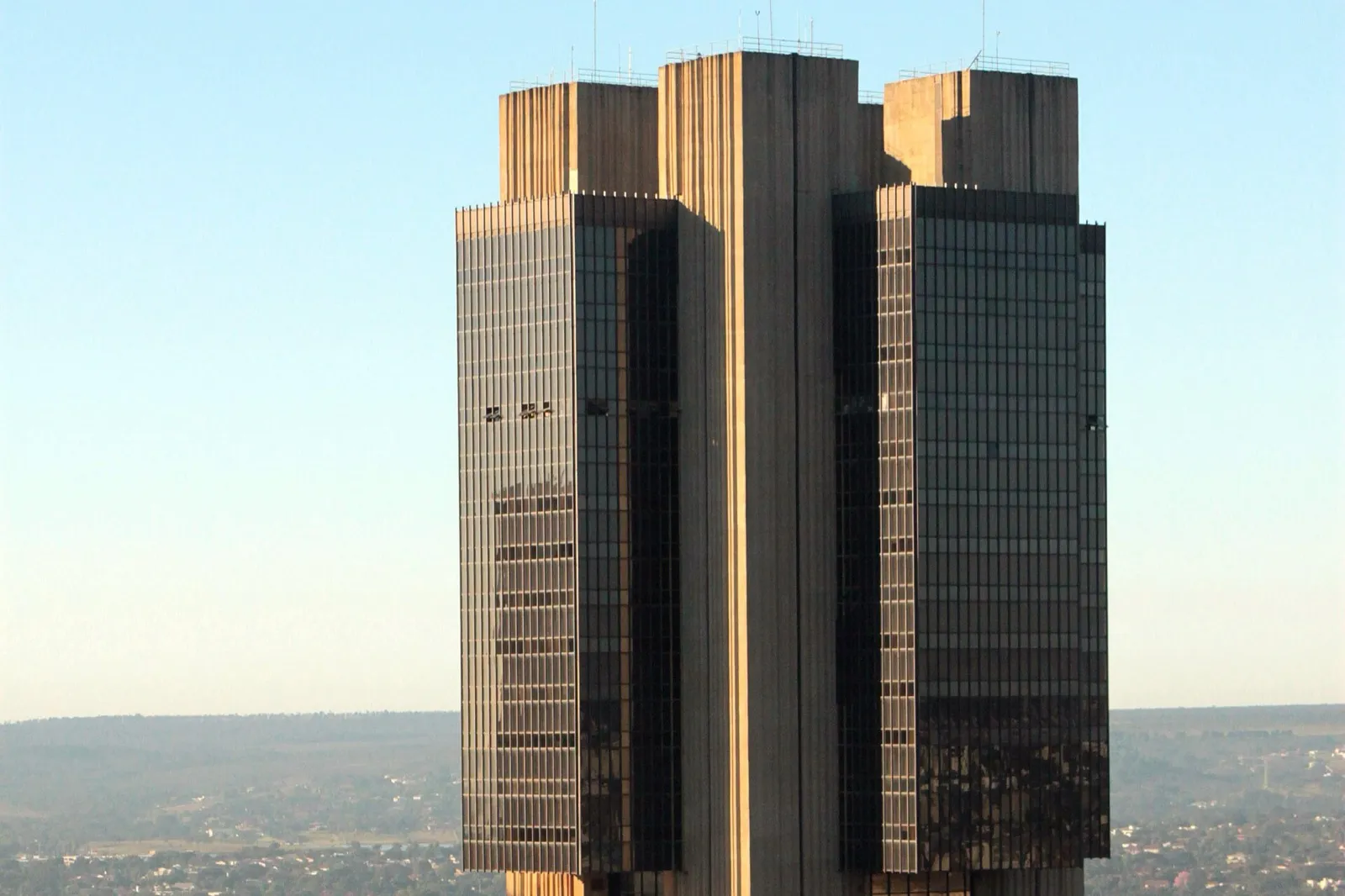 Torre de concreto e vidro escuro da sede do Banco Central do Brasil, em Brasília, sob céu azul, com as coberturas do prédio enquadradas.