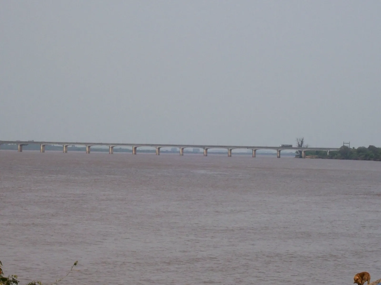 Longa ponte de concreto sobre o Rio Uruguai, com um caminhão atravessando, sob céu cinza e água barrenta.