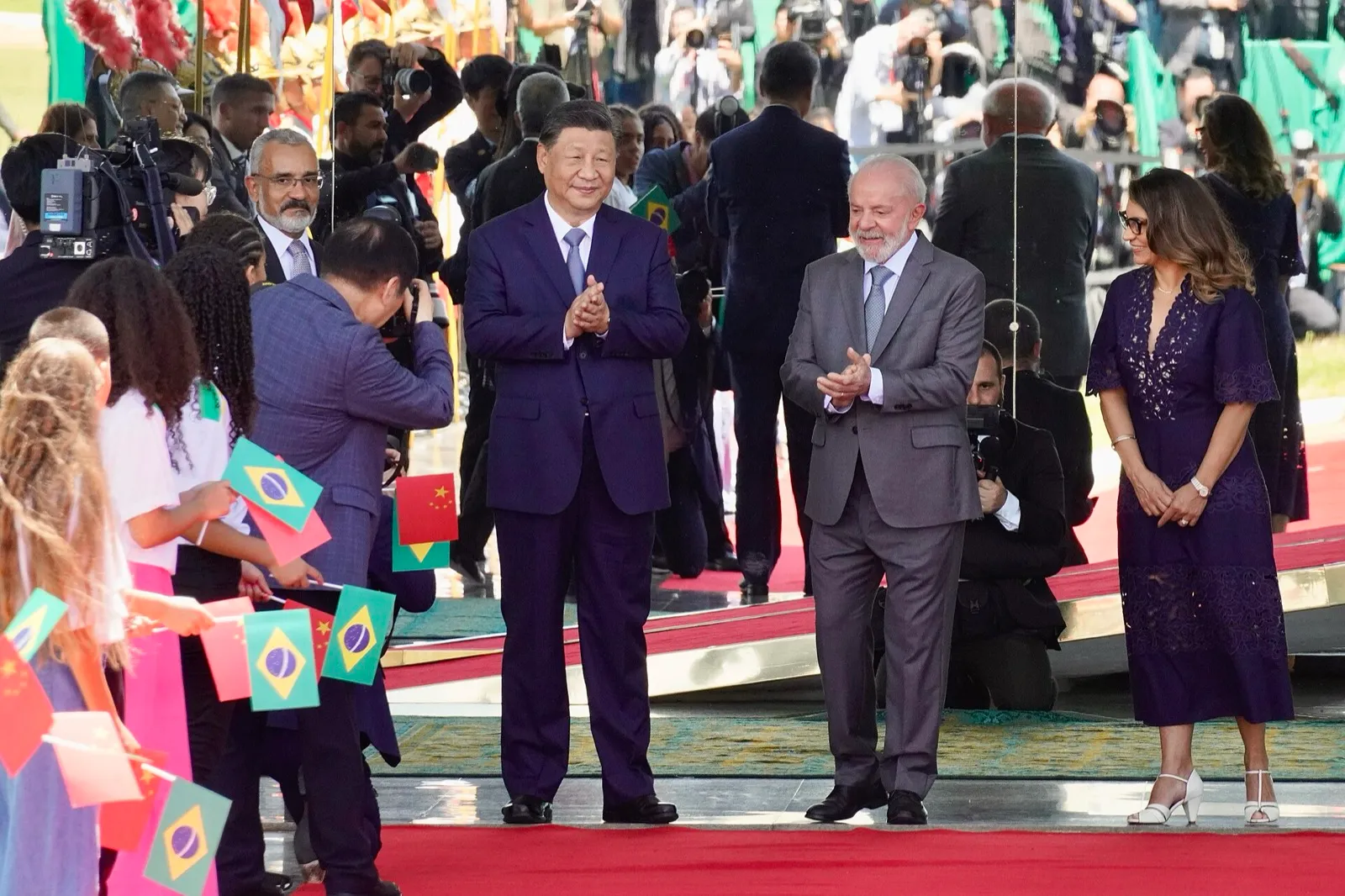 Xi Jinping e Lula aplaudem lado a lado sobre um tapete vermelho, diante de crianças com bandeirinhas do Brasil e da China.
