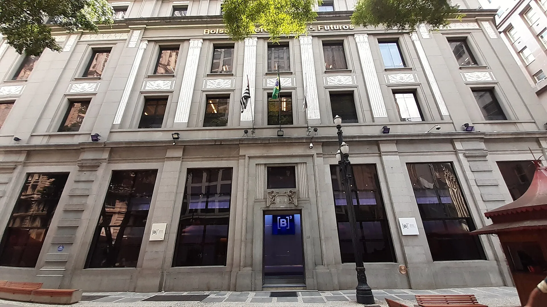 Fachada de pedra do edifício histórico da antiga Bolsa de Mercadorias & Futuros, com a porta de entrada iluminada pelo logotipo azul da B3.