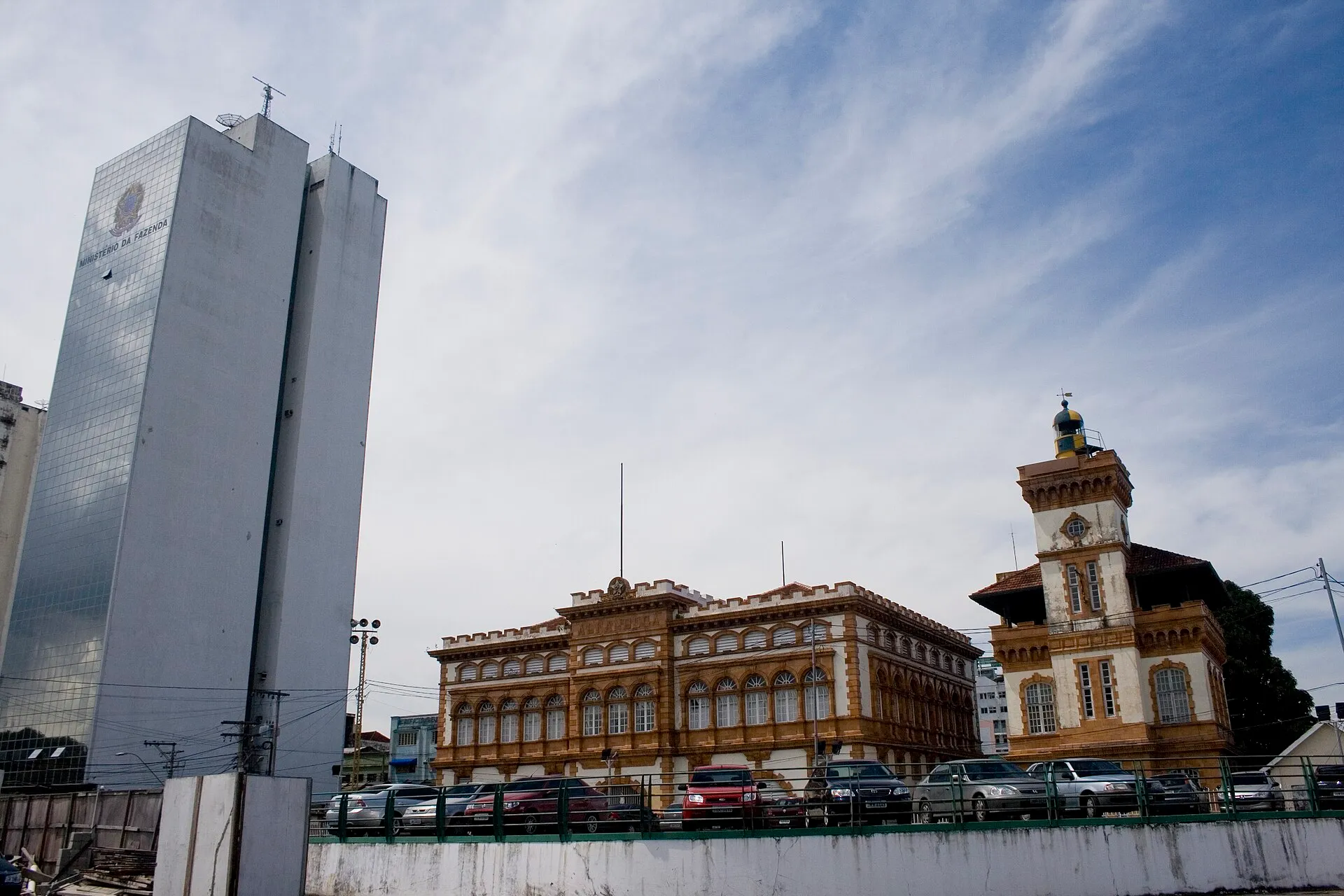Prédio em tons de ocre da Alfândega de Manaus, com ameias e torre, ao lado de um prédio espelhado do Ministério da Fazenda.