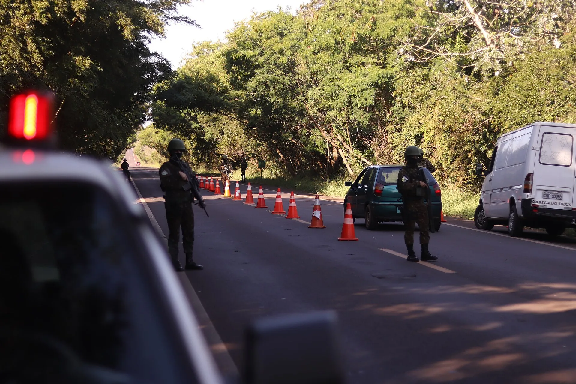 Militares do Exército com fuzis em posto de bloqueio numa estrada cercada de árvores, com cones laranja e carros parados para revista.