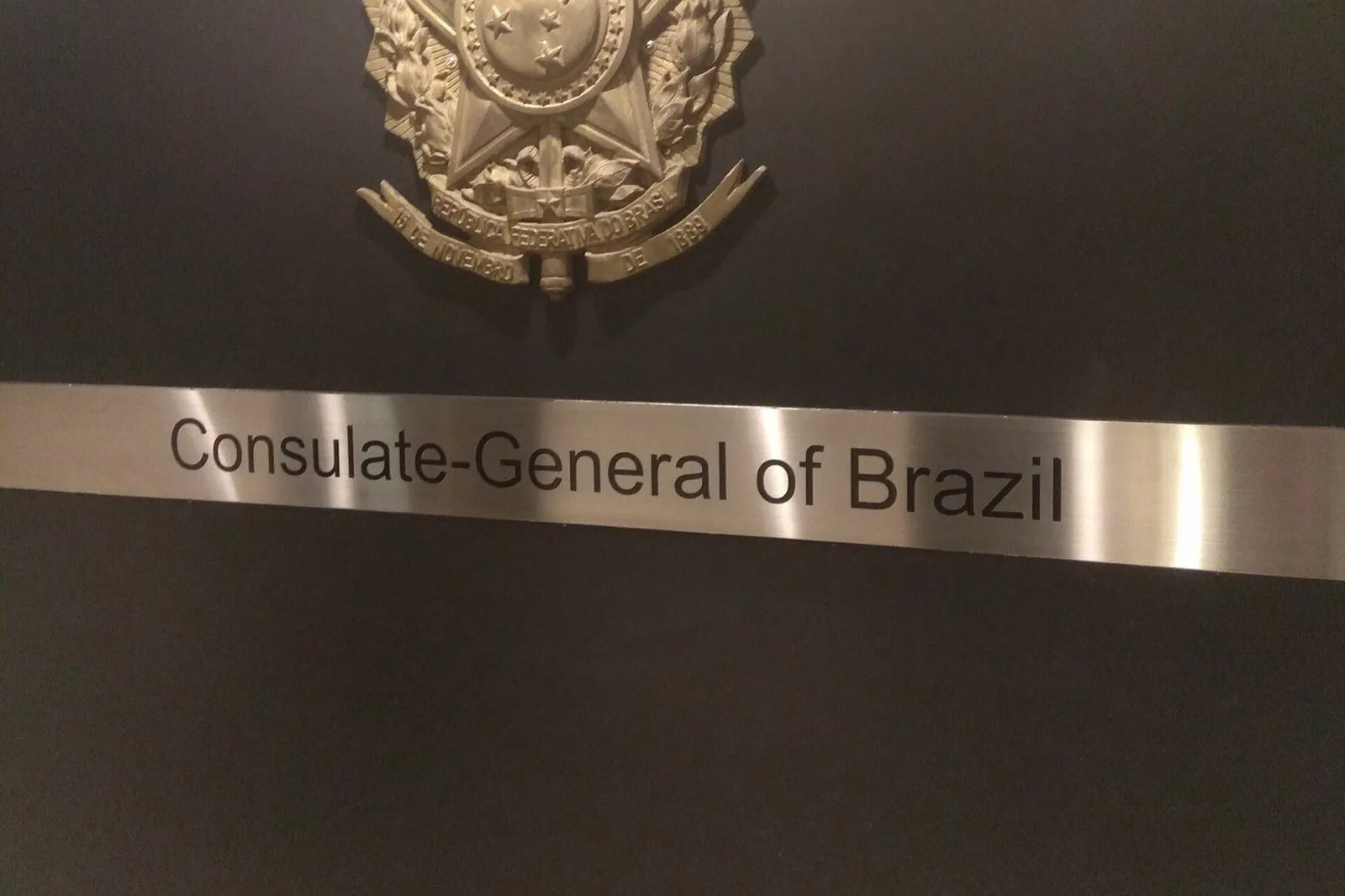 Placa de metal com o brasão da República Federativa do Brasil e a inscrição Consulate-General of Brazil, sobre parede escura.