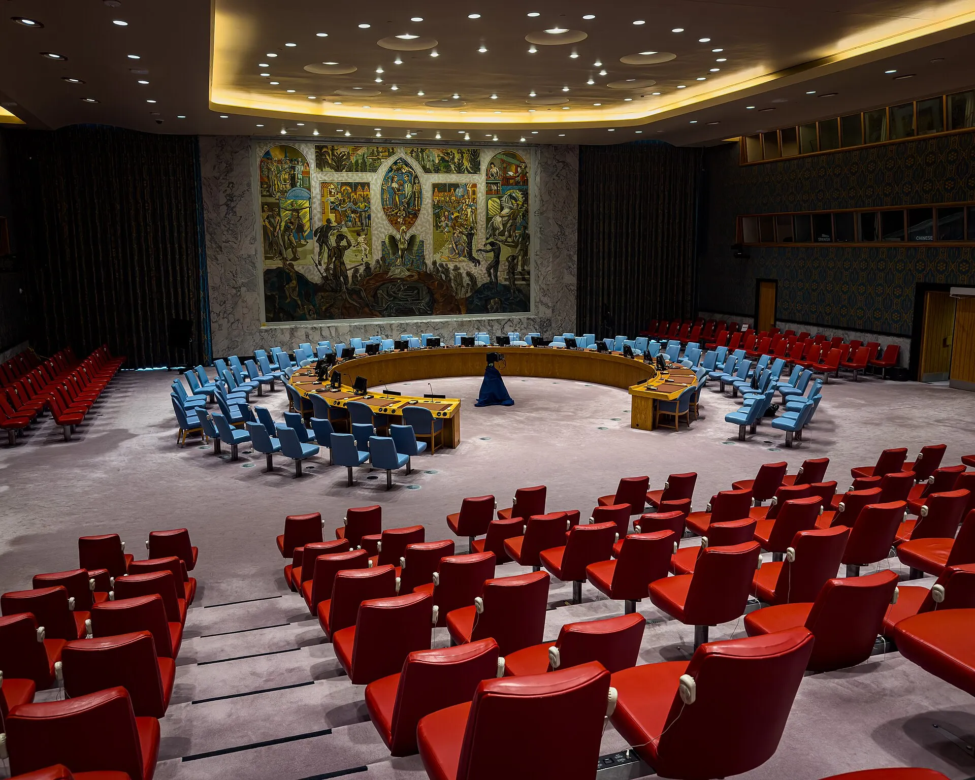 Plenário do Conselho de Segurança da ONU, vazio, com a mesa em ferradura, cadeiras azuis e vermelhas e um grande mural ao fundo.