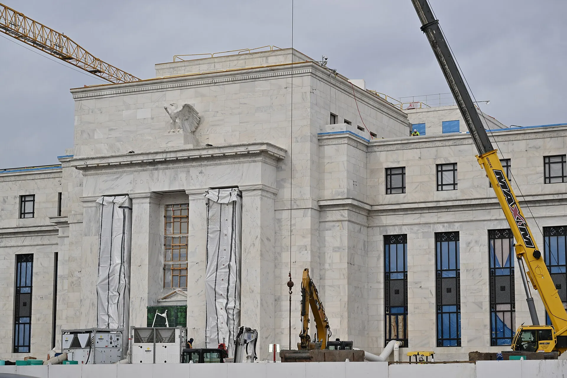 Fachada do prédio do Federal Reserve em Washington durante obras, com guindastes e colunas de mármore.