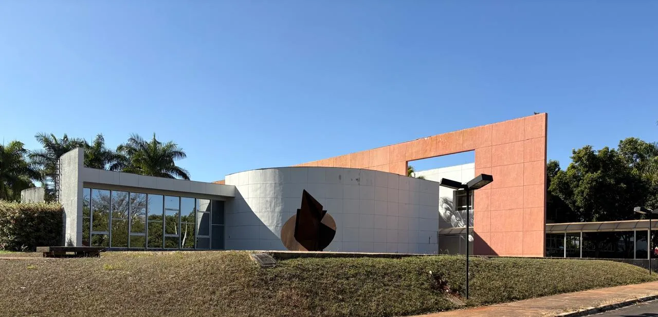 Prédio do Instituto Rio Branco, em Brasília, com fachada de concreto em tons claros e laranja, uma escultura metálica escura à frente e céu azul.