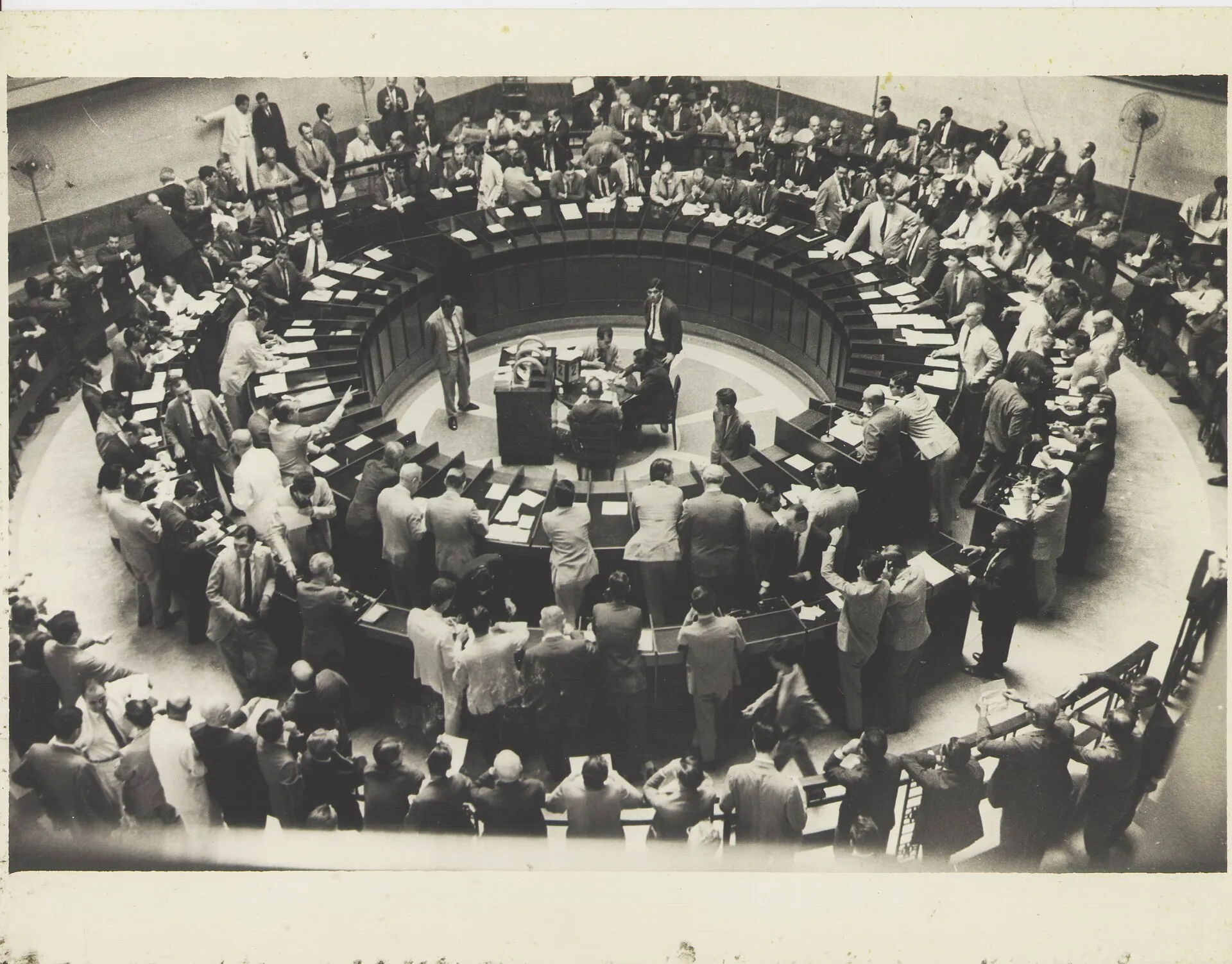 Foto em preto e branco de um pregão de bolsa, com dezenas de homens de paletó em volta de uma mesa circular e papéis sobre as bancadas.