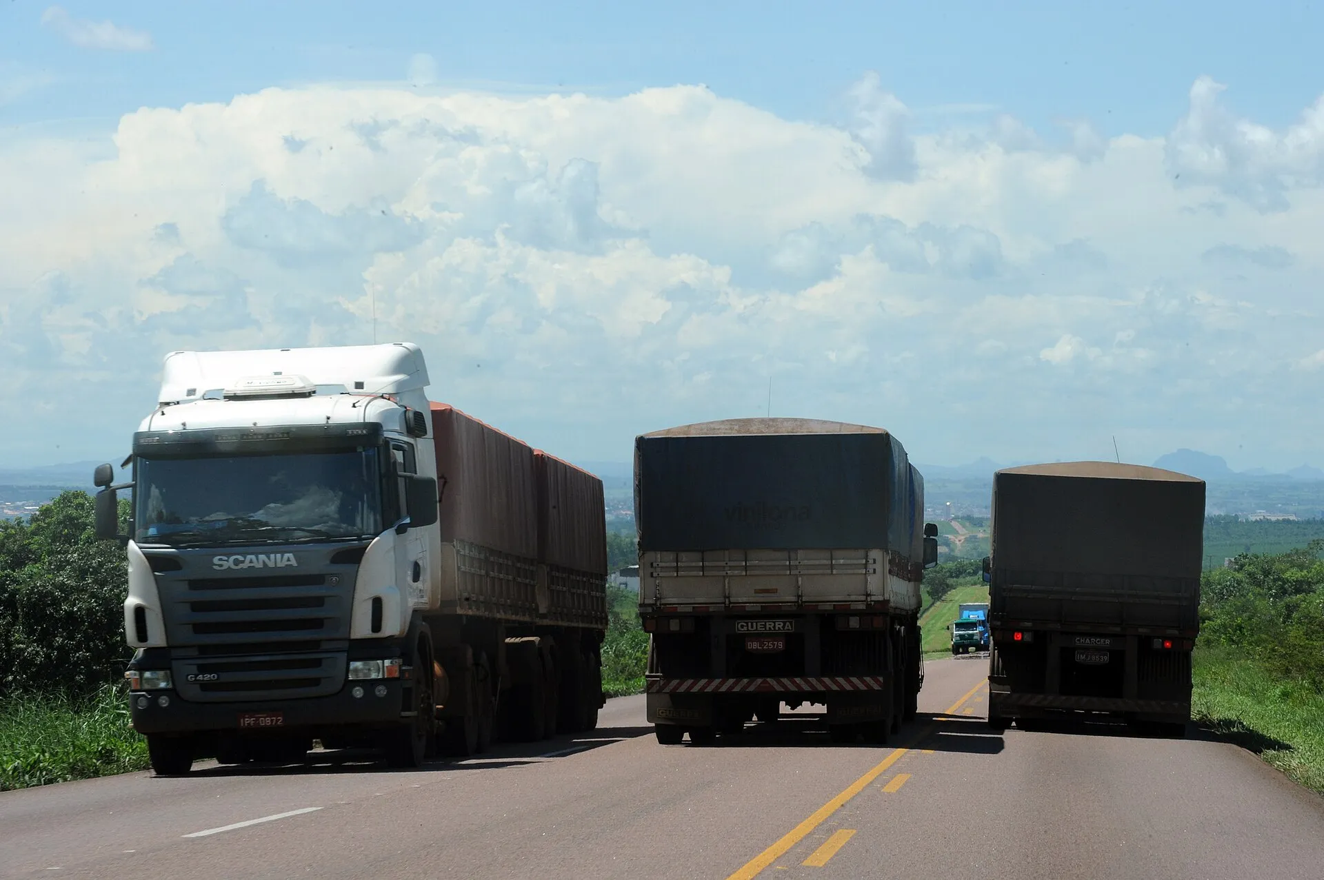 Três caminhões graneleiros com carga coberta por lona seguem por uma rodovia de pista dupla, sob céu azul com nuvens grandes.