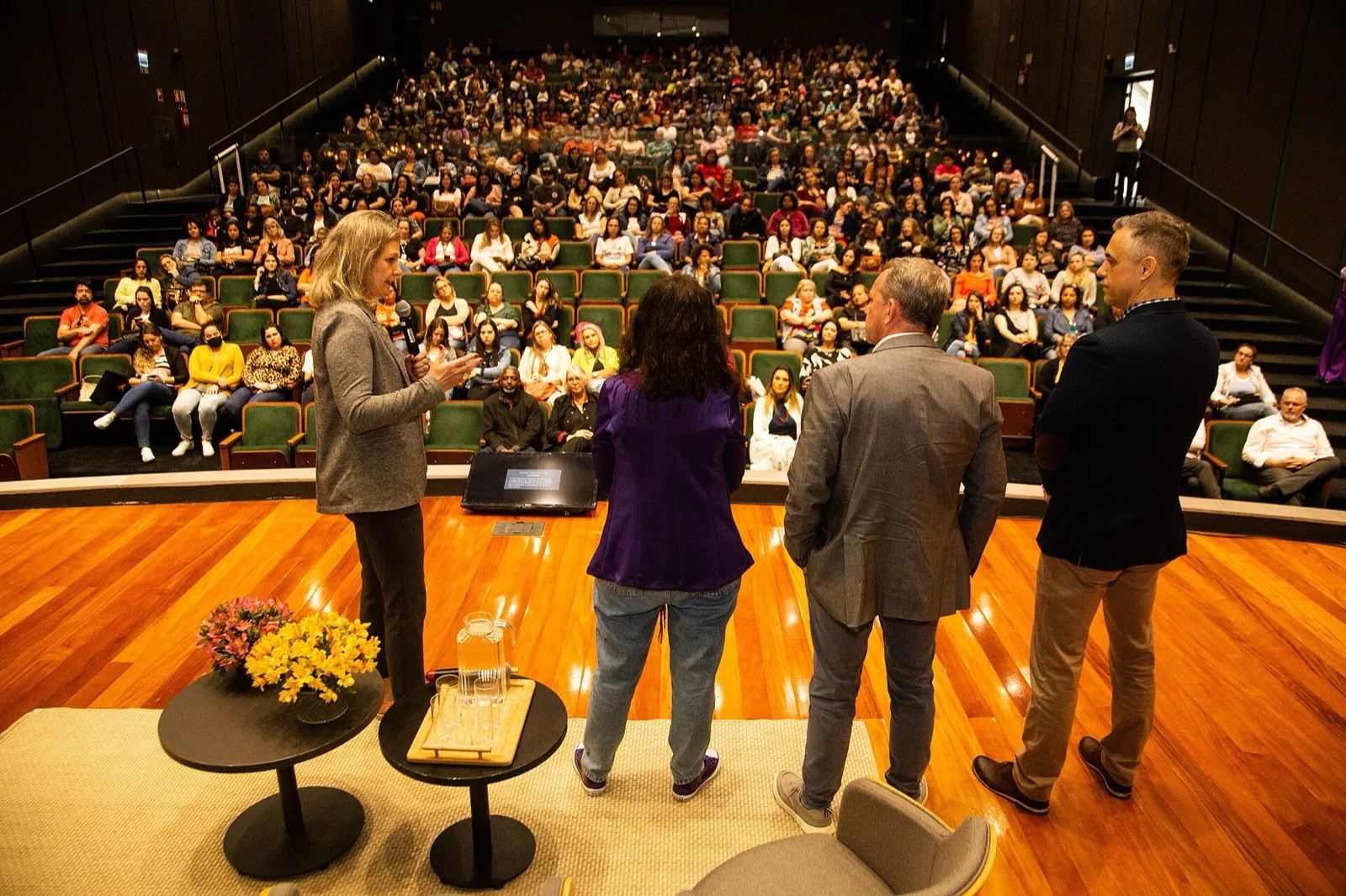 Uma mulher com microfone fala a uma plateia lotada de professores em um auditório, enquanto três pessoas observam do palco.