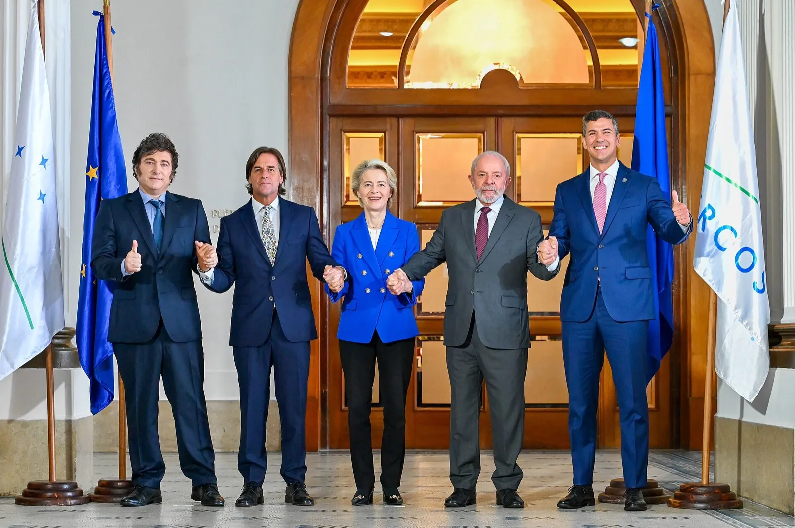 Cinco líderes de mãos dadas, entre bandeiras da União Europeia e do Mercosul, diante de uma porta de madeira.