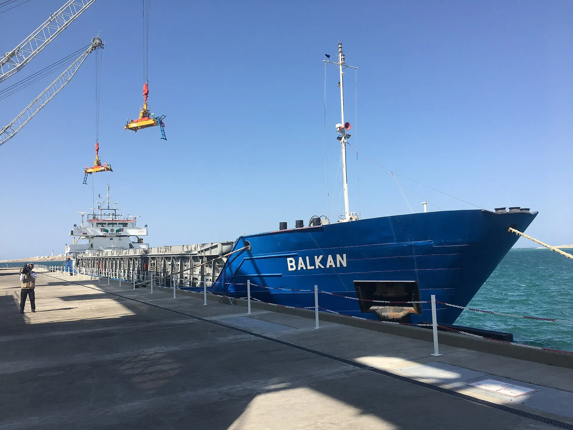 Navio azul chamado Balkan atracado em um cais de carregamento, com guindastes no alto e mar azul-turquesa ao fundo, sob céu claro.