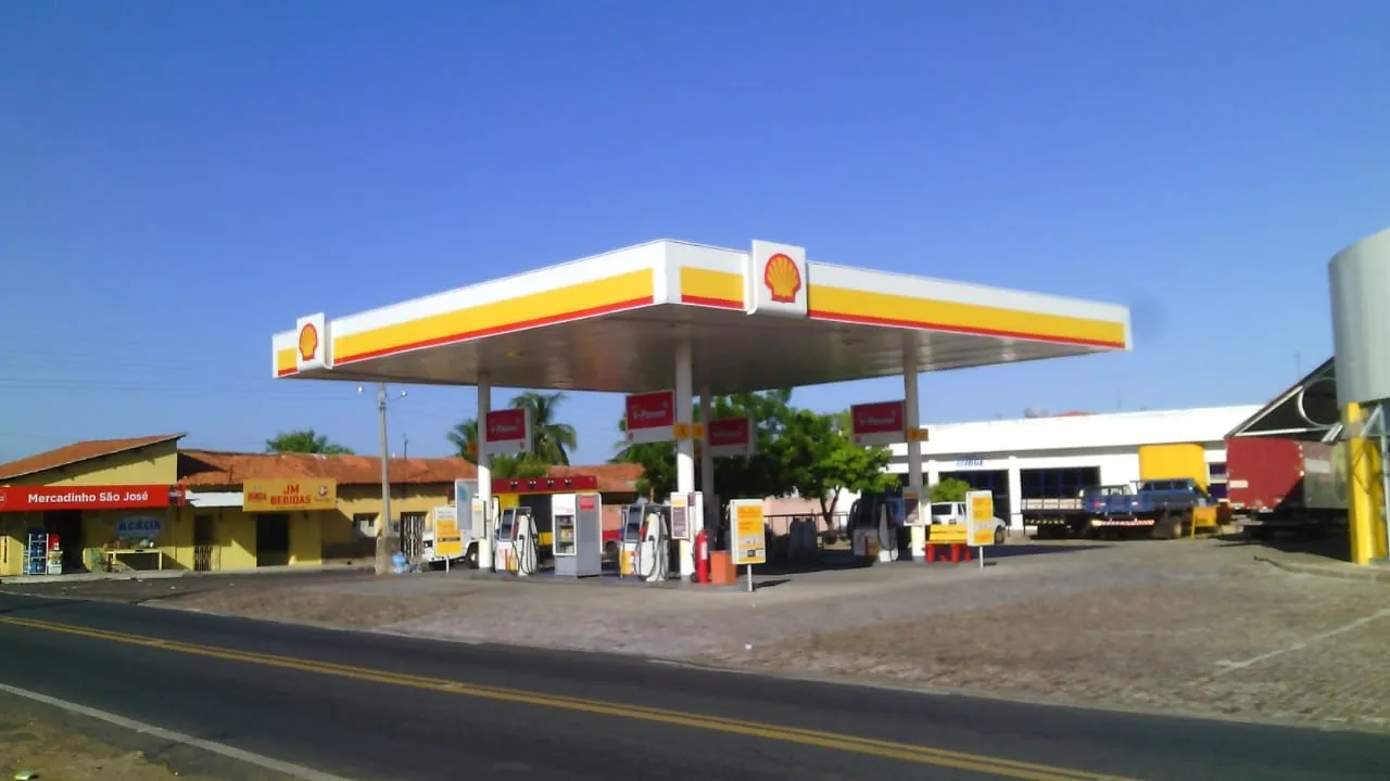 Posto de combustíveis à margem de uma rodovia, com bombas, caminhões e cobertura nas cores amarelo e vermelho.