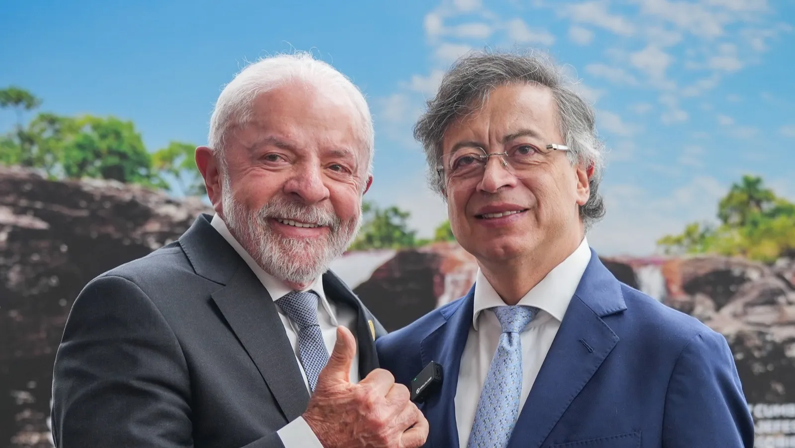 Lula, de terno cinza, faz sinal de positivo ao lado de Gustavo Petro, de terno azul, diante de um painel com cachoeira e céu azul.