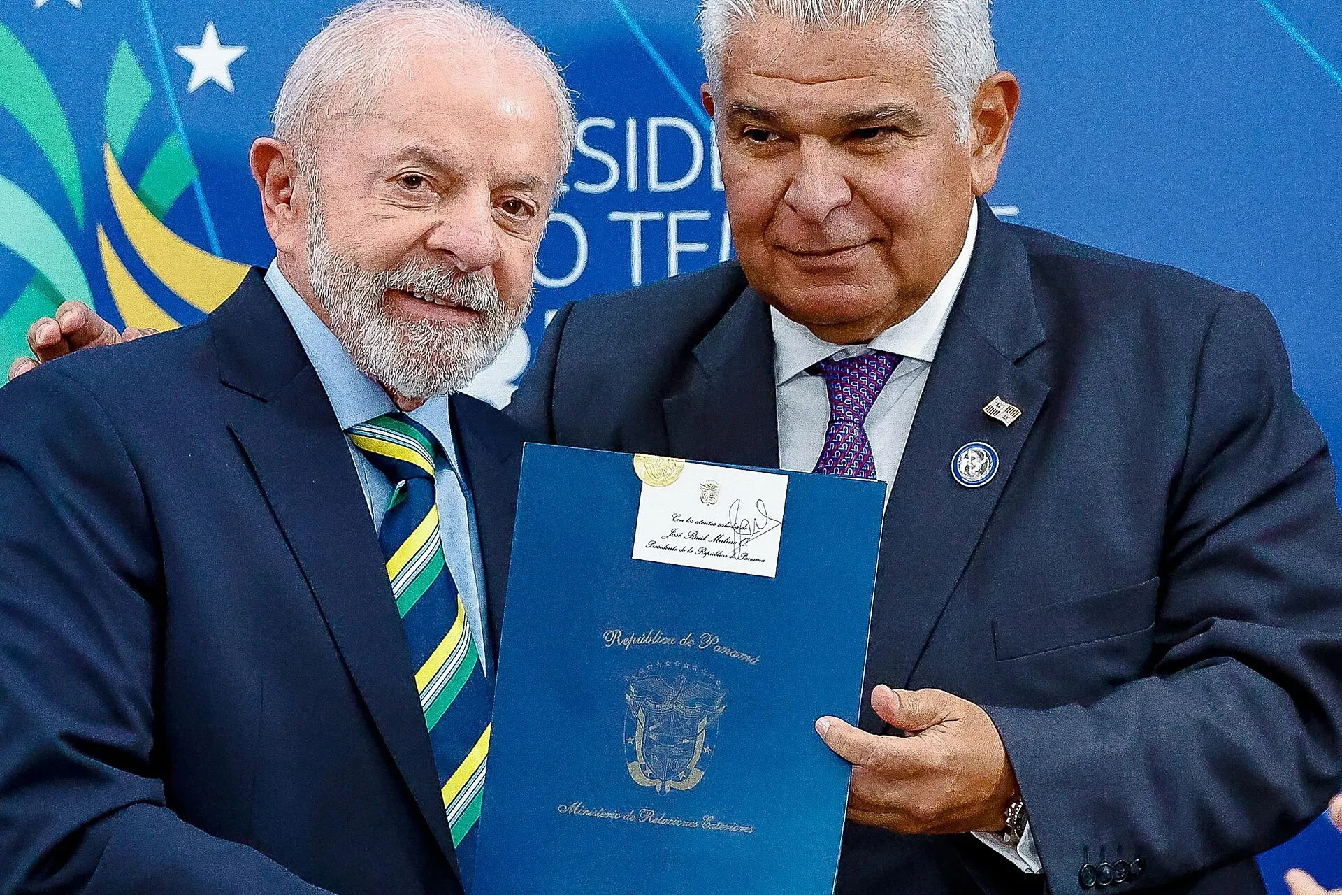 Lula, à esquerda, e o presidente do Panamá, José Raúl Mulino, à direita, seguram juntos uma pasta azul do Ministério de Relações Exteriores do Panamá; sobre a mesa, bandeiras do Brasil e do Mercosul.