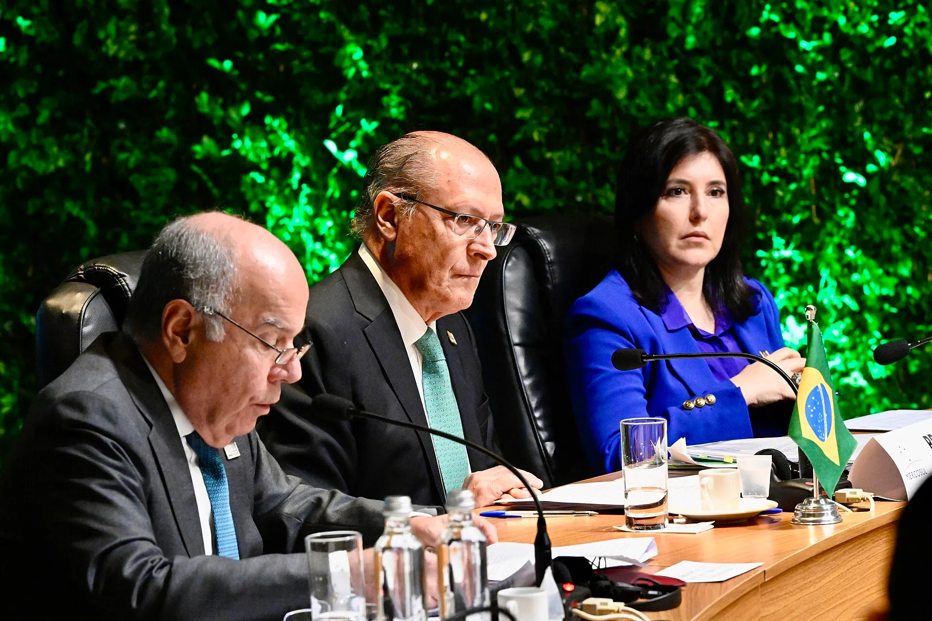 Três autoridades brasileiras sentadas à mesa de uma reunião do Mercosul, diante de uma parede de folhagem iluminada em verde, com a bandeira do Brasil em primeiro plano.