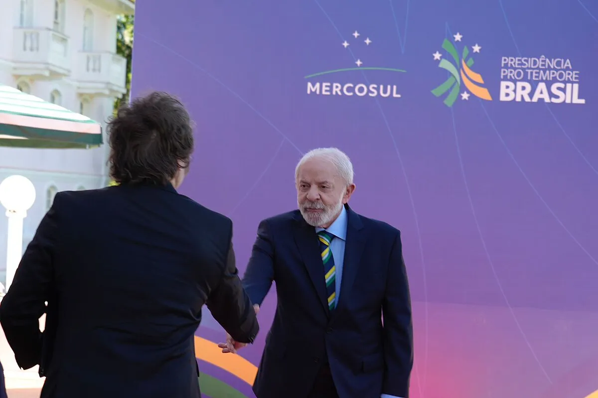 Lula, de terno azul-marinho e gravata listrada, cumprimenta Javier Milei, de costas, diante de painel roxo com os logos do Mercosul e da Presidência Pro Tempore do Brasil.