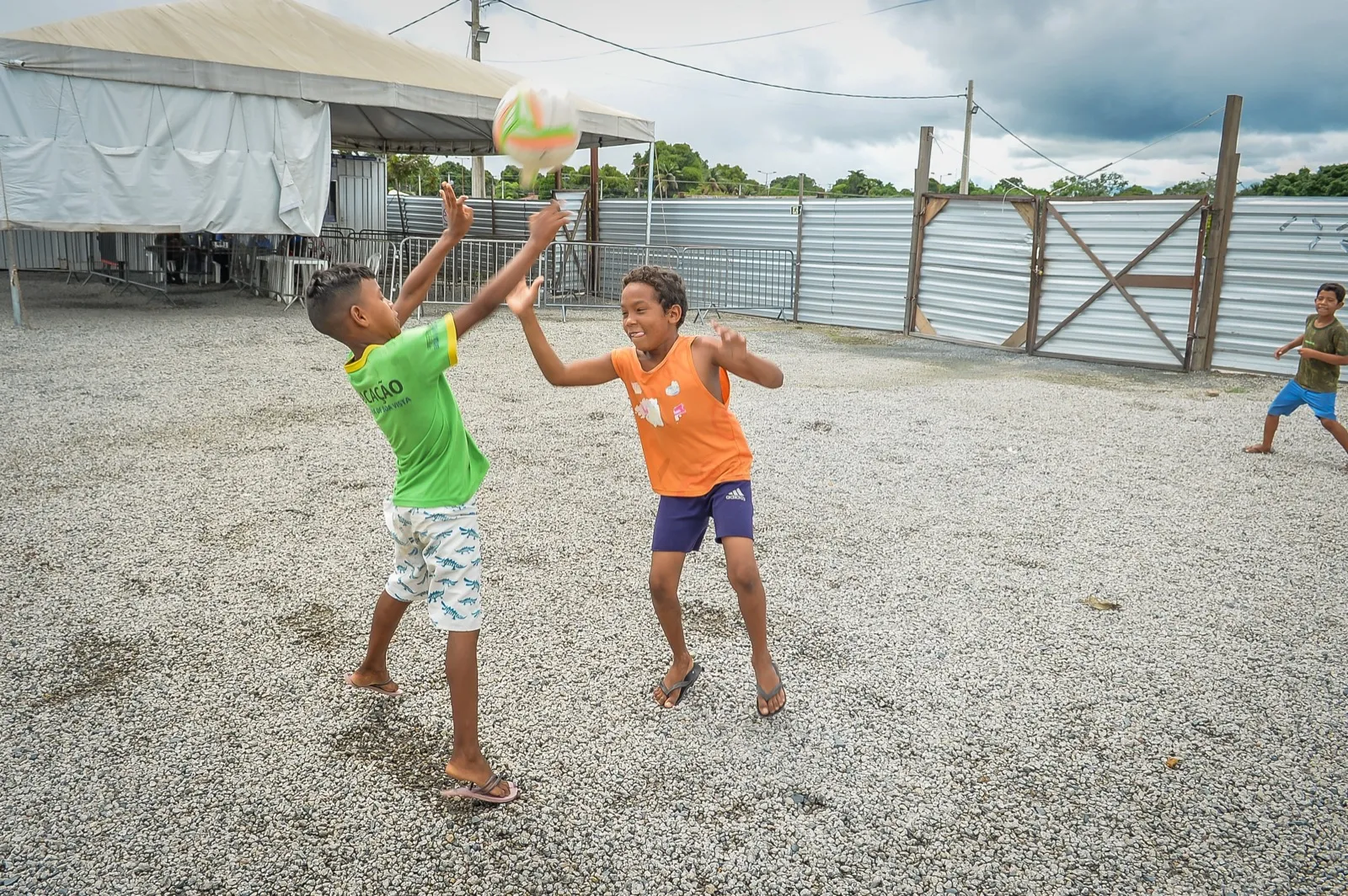 Dois meninos disputam uma bola em um pátio de terra com tenda branca e cercas de metal, em um abrigo da Operação Acolhida em Boa Vista.