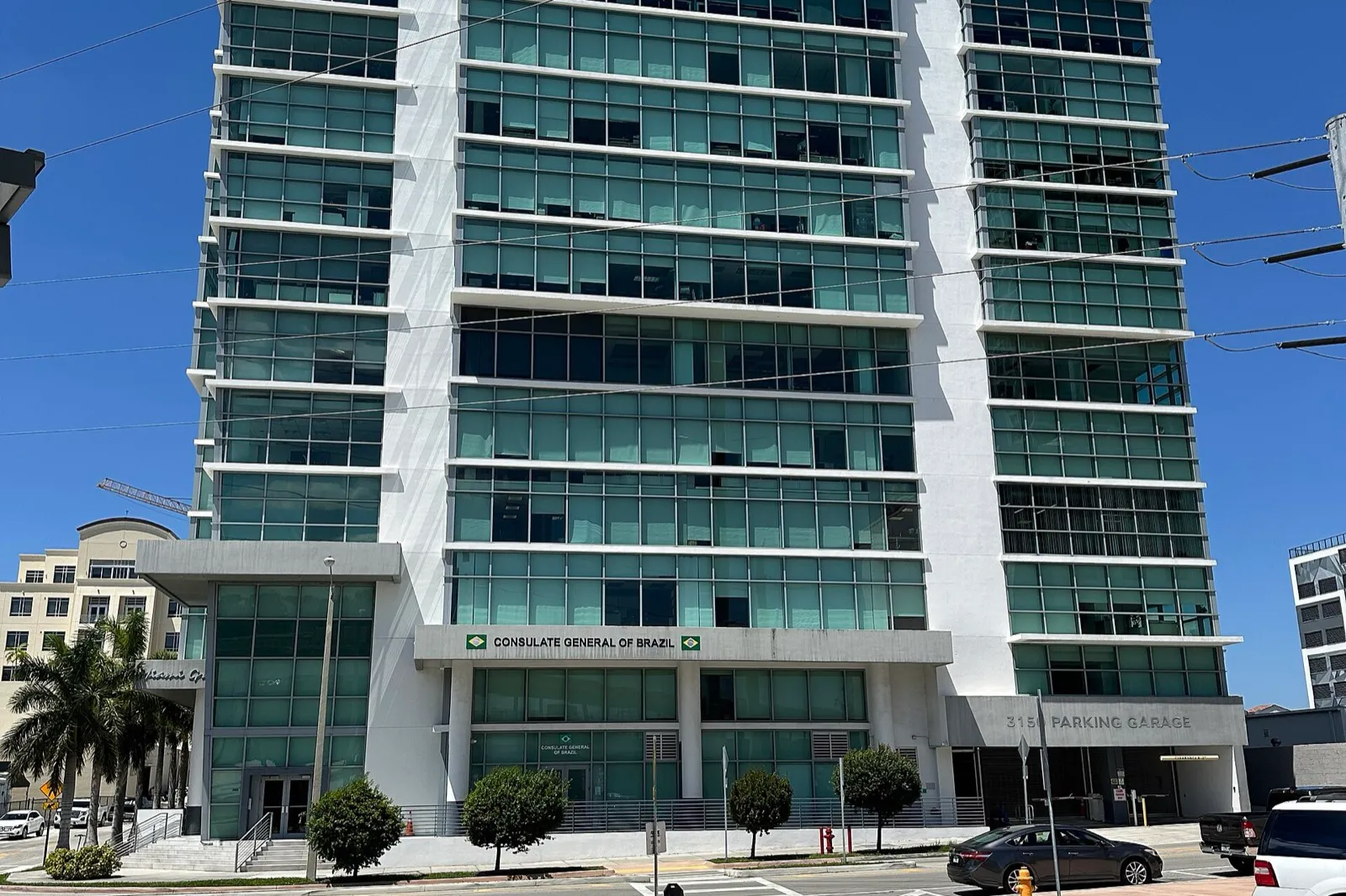 Prédio branco e envidraçado com a placa Consulate General of Brazil, com bandeiras do Brasil, sob céu azul.