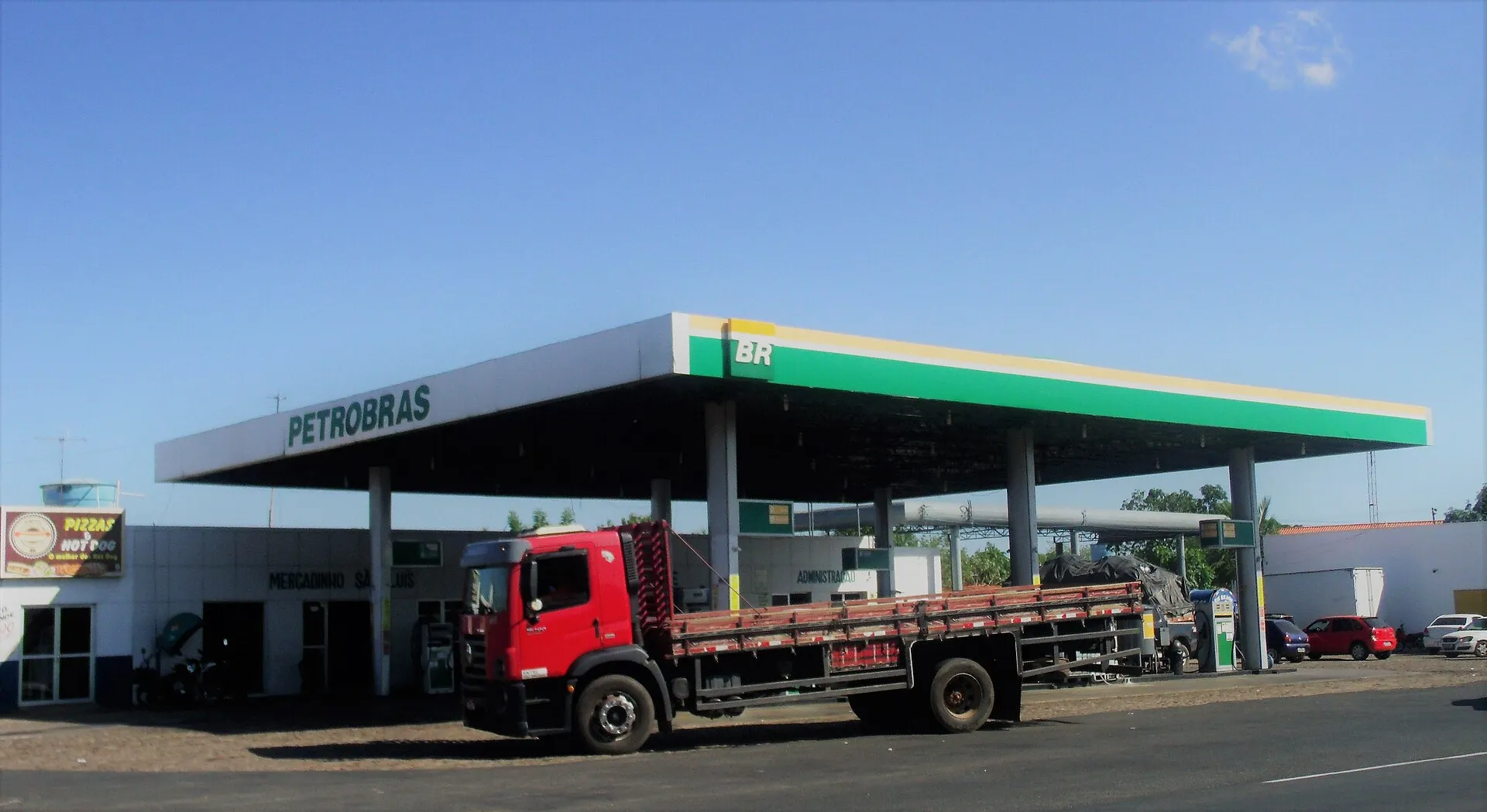 Posto de combustível da Petrobras, com cobertura verde e amarela e a marca BR, sob céu azul. Um caminhão vermelho de carroceria aberta está parado na frente.