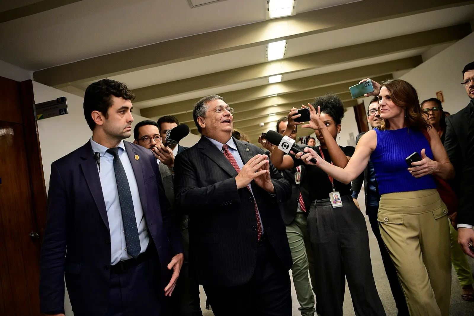 Flávio Dino, de terno escuro e gravata vermelha, fala com jornalistas em um corredor do Senado Federal.