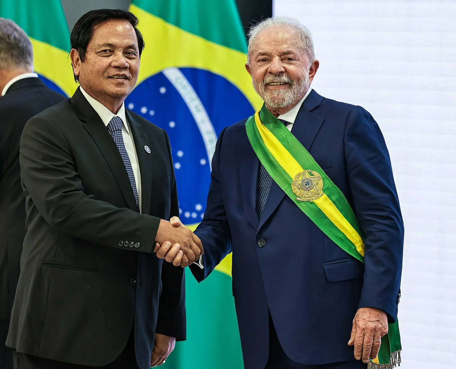 Lula, de terno azul-marinho e faixa presidencial verde e amarela, aperta a mão de um homem de terno escuro diante de uma bandeira do Brasil, no Palácio do Planalto.