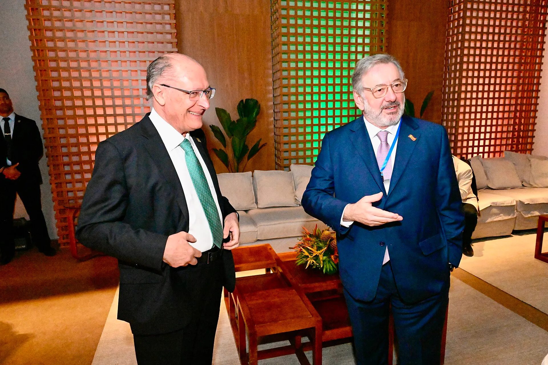 Dois homens de terno conversam em um salão com treliças de madeira iluminadas em verde e laranja; o da esquerda sorri e o da direita gesticula.
