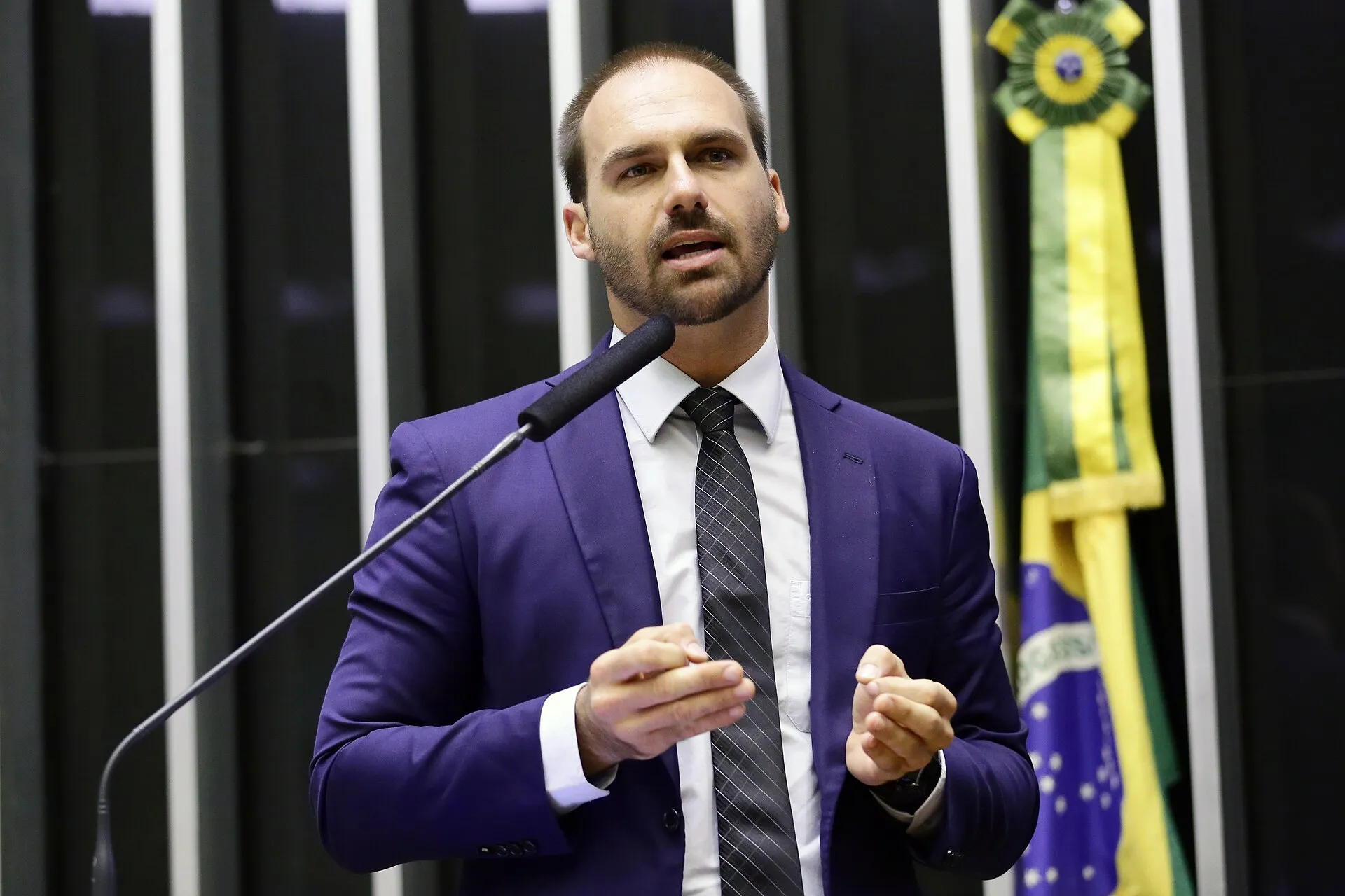 Homem de terno azul e gravata discursa ao microfone no plenário da Câmara dos Deputados, com a bandeira do Brasil ao fundo.