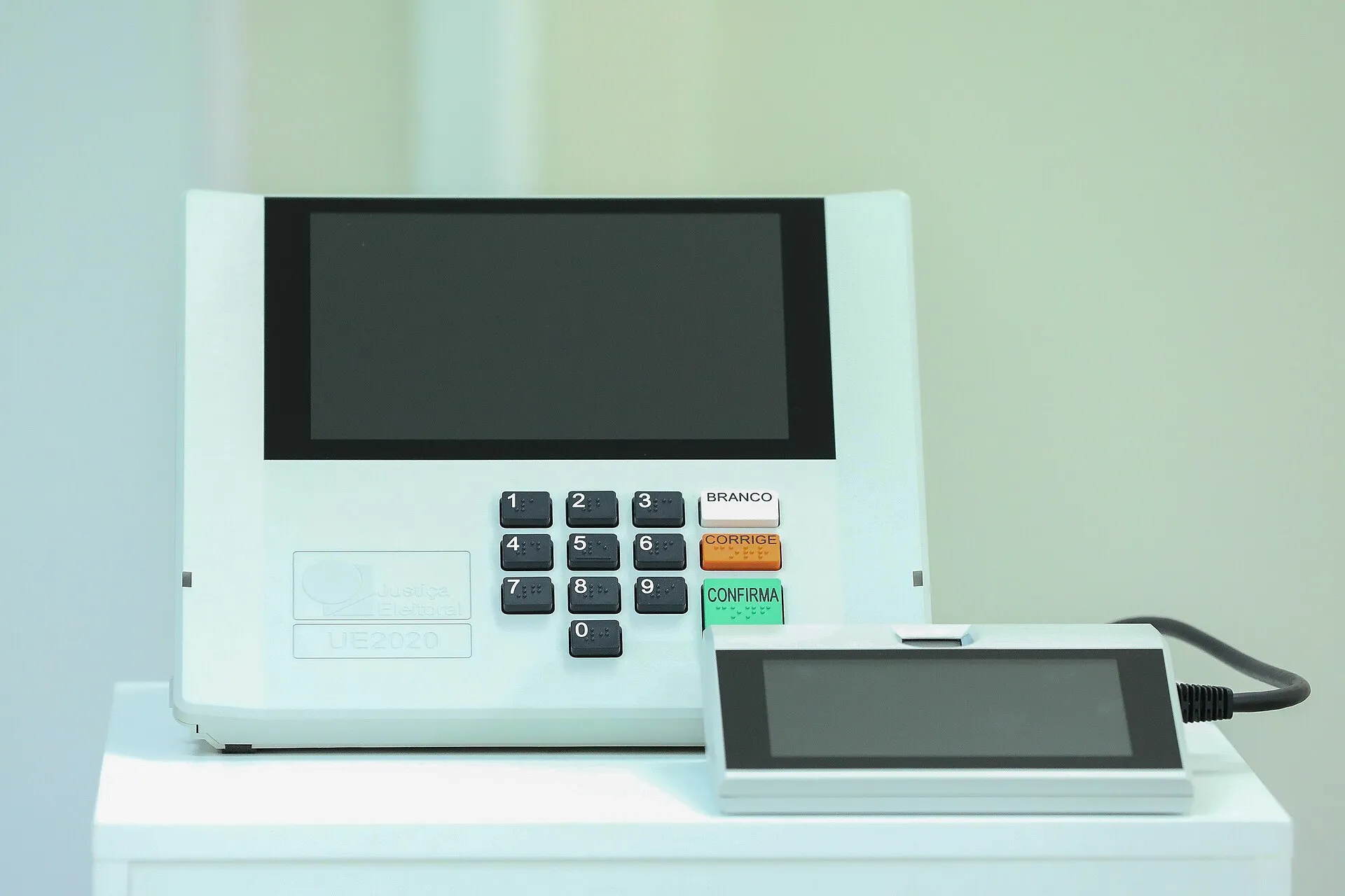 Urna eletrônica modelo UE2020, da Justiça Eleitoral, com a tela apagada, teclado numérico e um aparelho menor ao lado, sobre uma superfície branca.