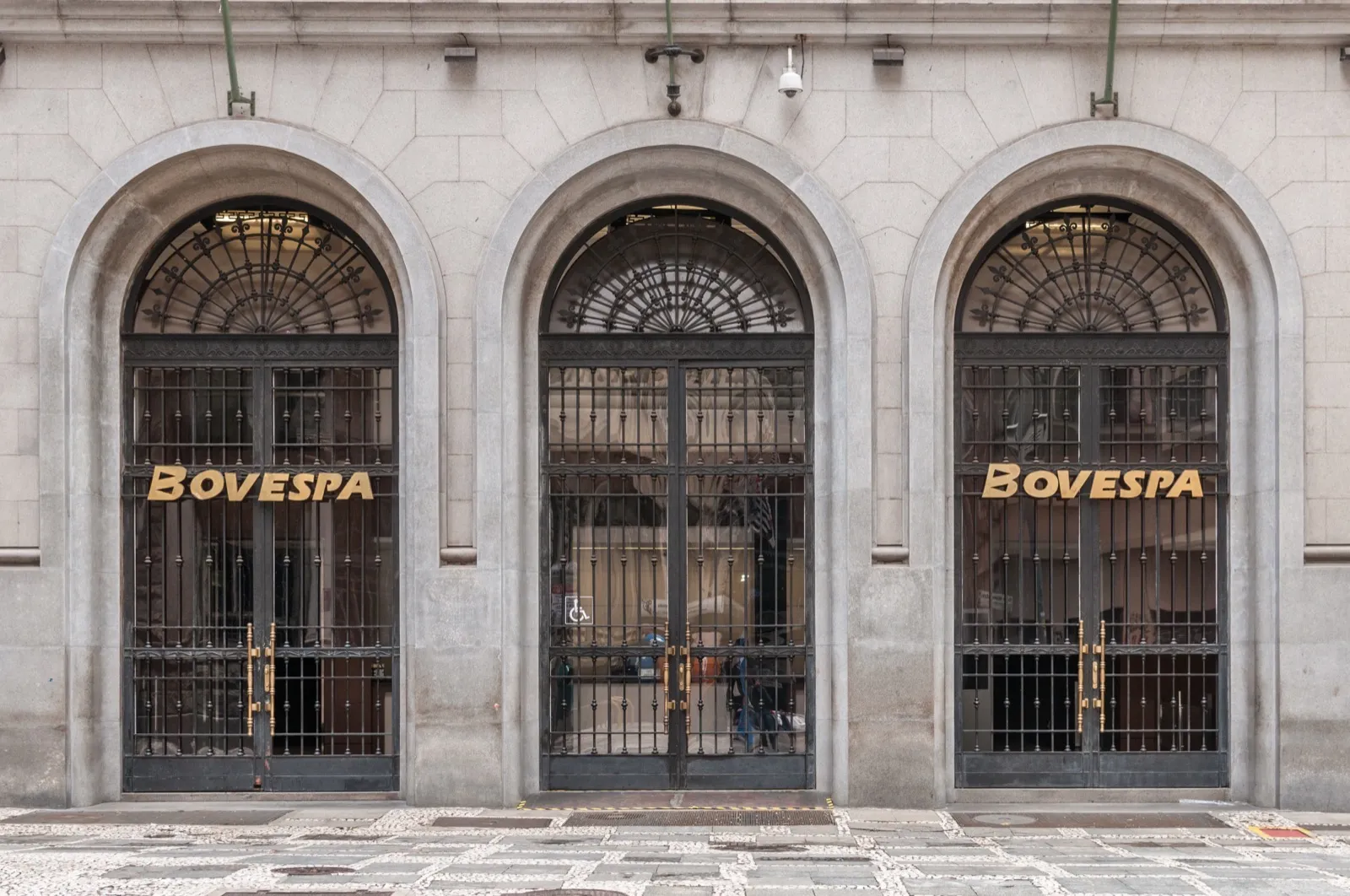 Entrada do prédio da Bovespa, em São Paulo, com três portas de ferro sob arcos de pedra e as letras BOVESPA em dourado.