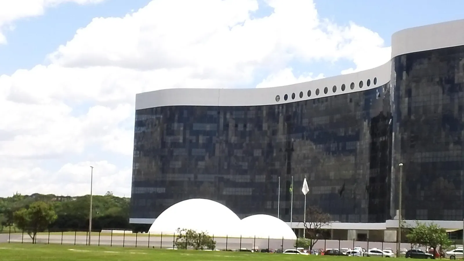 Edifício-sede do Tribunal Superior Eleitoral em Brasília, com fachada escura e espelhada, duas cúpulas brancas na frente e gramado verde.
