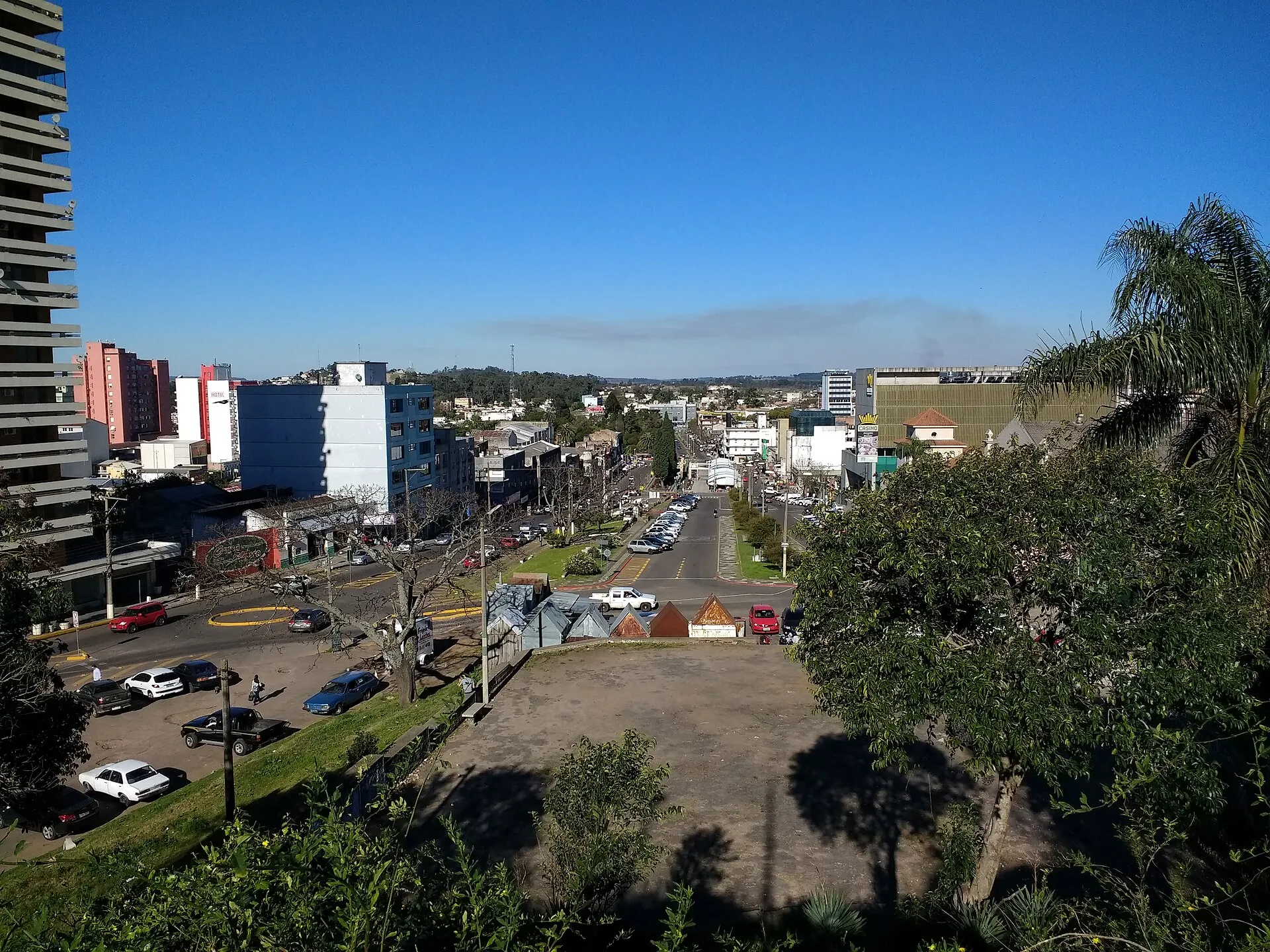 Vista elevada de uma avenida larga, com carros estacionados, prédios baixos e altos e céu azul, em uma cidade de fronteira entre o Brasil e o Uruguai.
