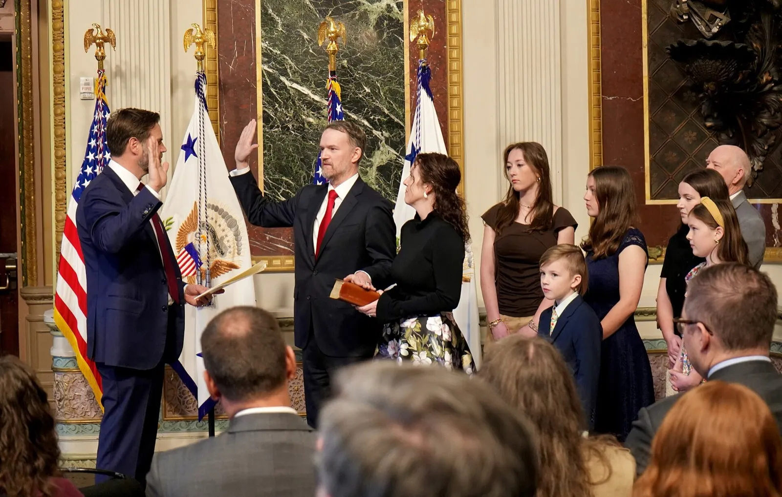 Homem de terno escuro levanta a mão direita em juramento, diante de bandeiras dos Estados Unidos, em cerimônia com familiares e público.