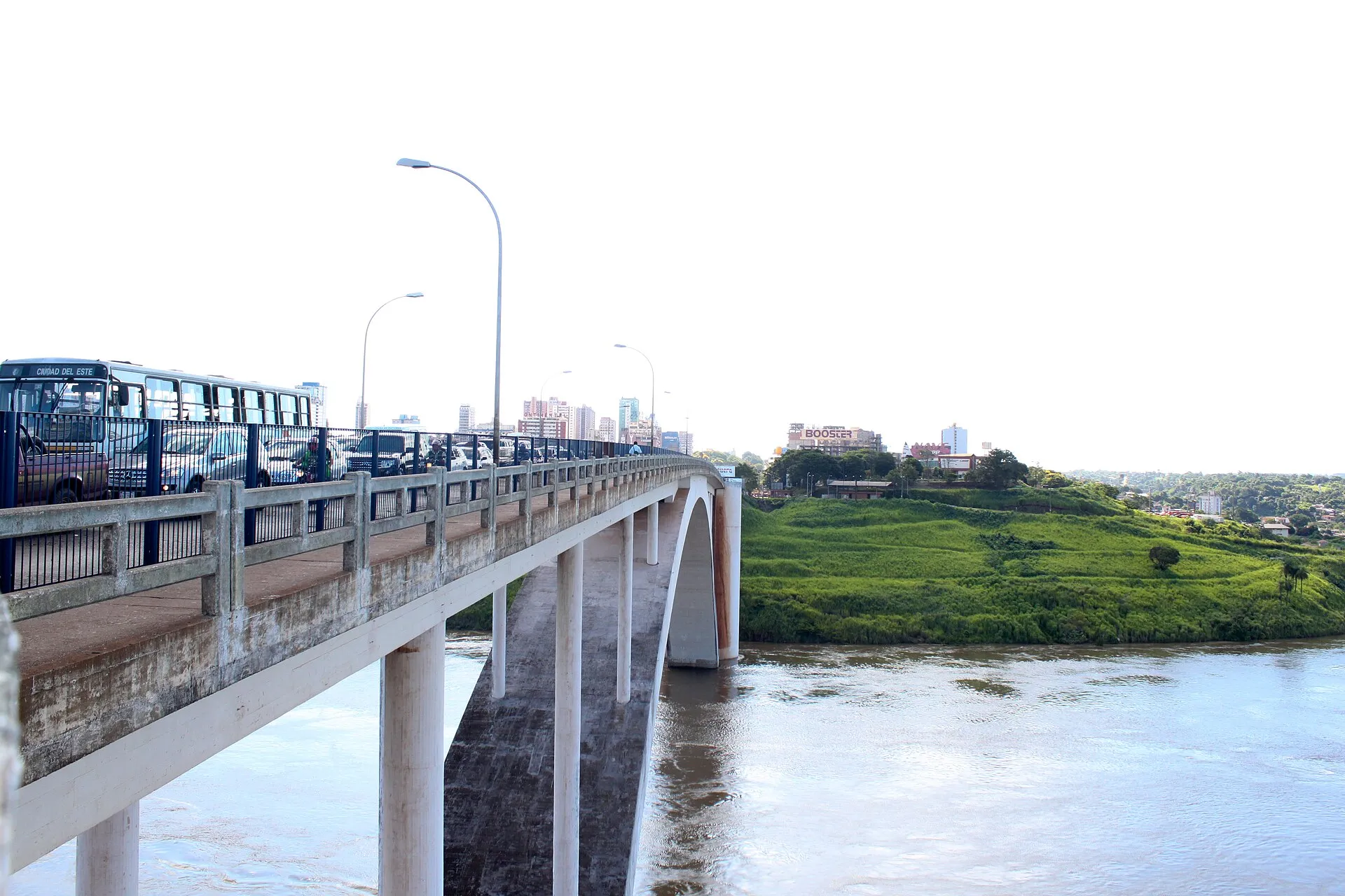 Ponte da Amizade vista de cima, com ônibus e carros na pista, o rio Paraná embaixo e a cidade ao fundo.