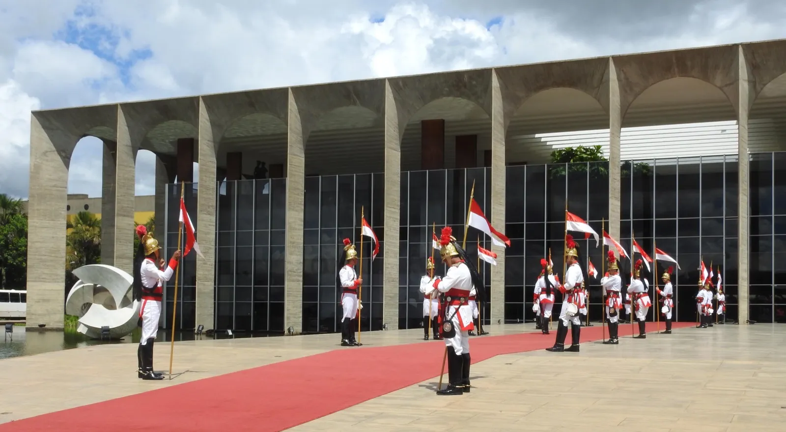 Fachada do Palácio do Itamaraty, em Brasília, com colunas de concreto, painéis de vidro escuro e guardas cerimoniais de uniforme branco e vermelho segurando bandeiras.